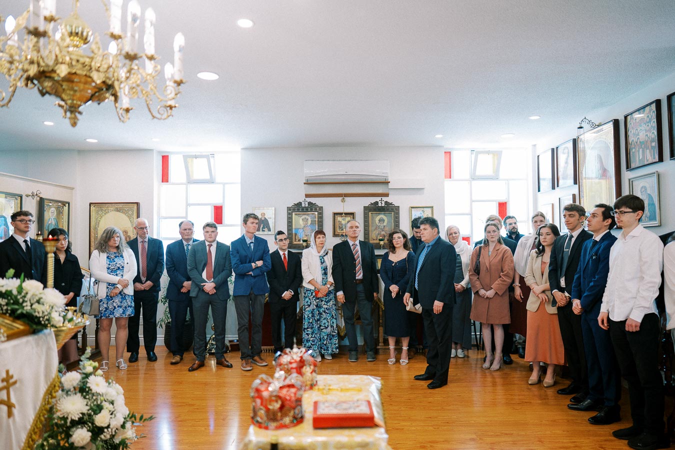 A group of formally dressed individuals stand together in a bright room during a religious ceremony, surrounded by iconography and floral decorations, with a gold chandelier overhead.