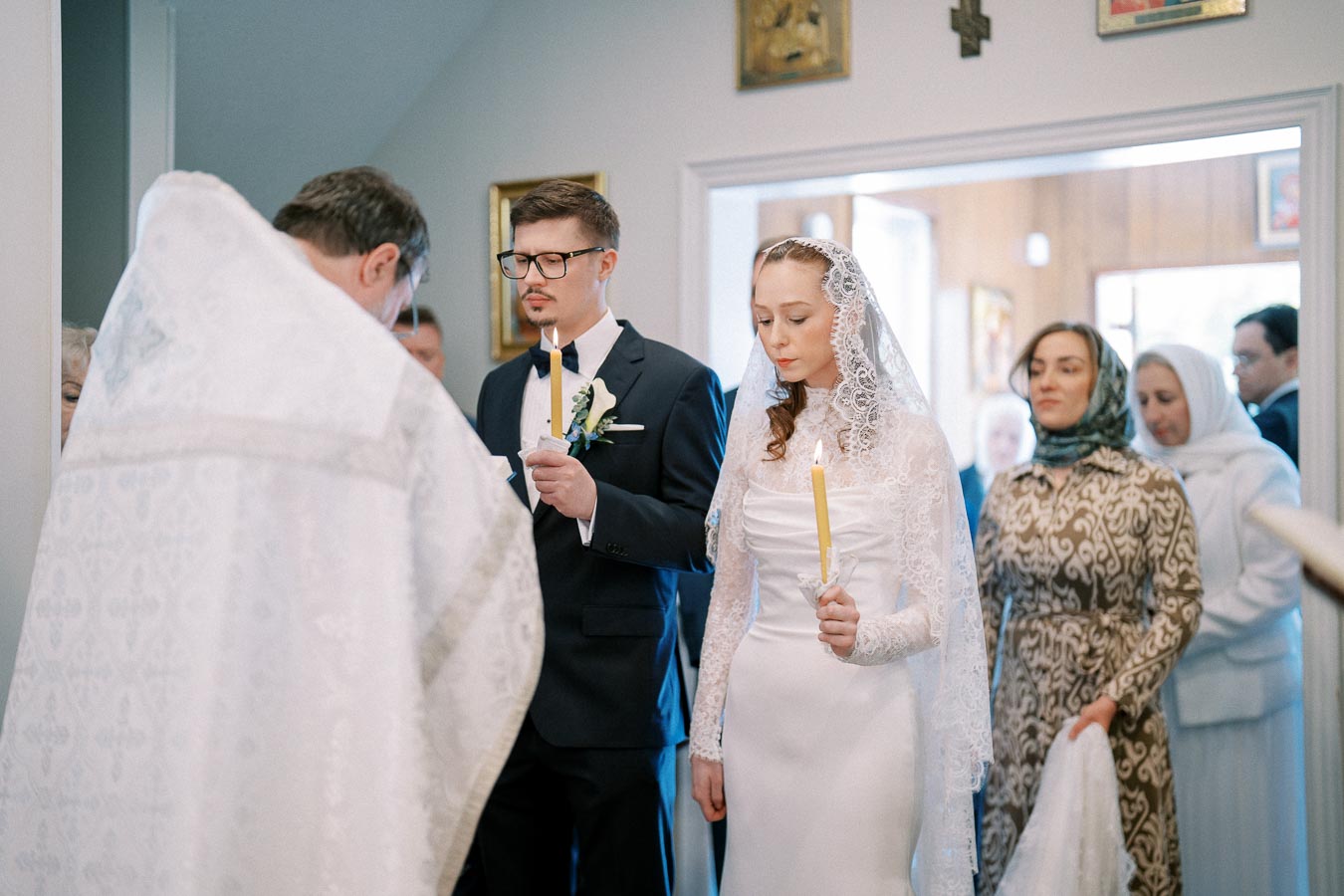 A bride and groom holding candles during an Orthodox wedding ceremony, with a priest conducting the ritual and attendees in the background, inside a decorated church space.