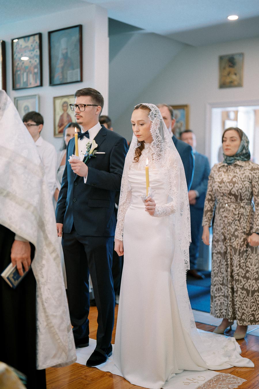 Orthodox wedding ceremony with bride and groom holding candles, dressed in traditional attire, surrounded by guests in a church setting adorned with religious icons.