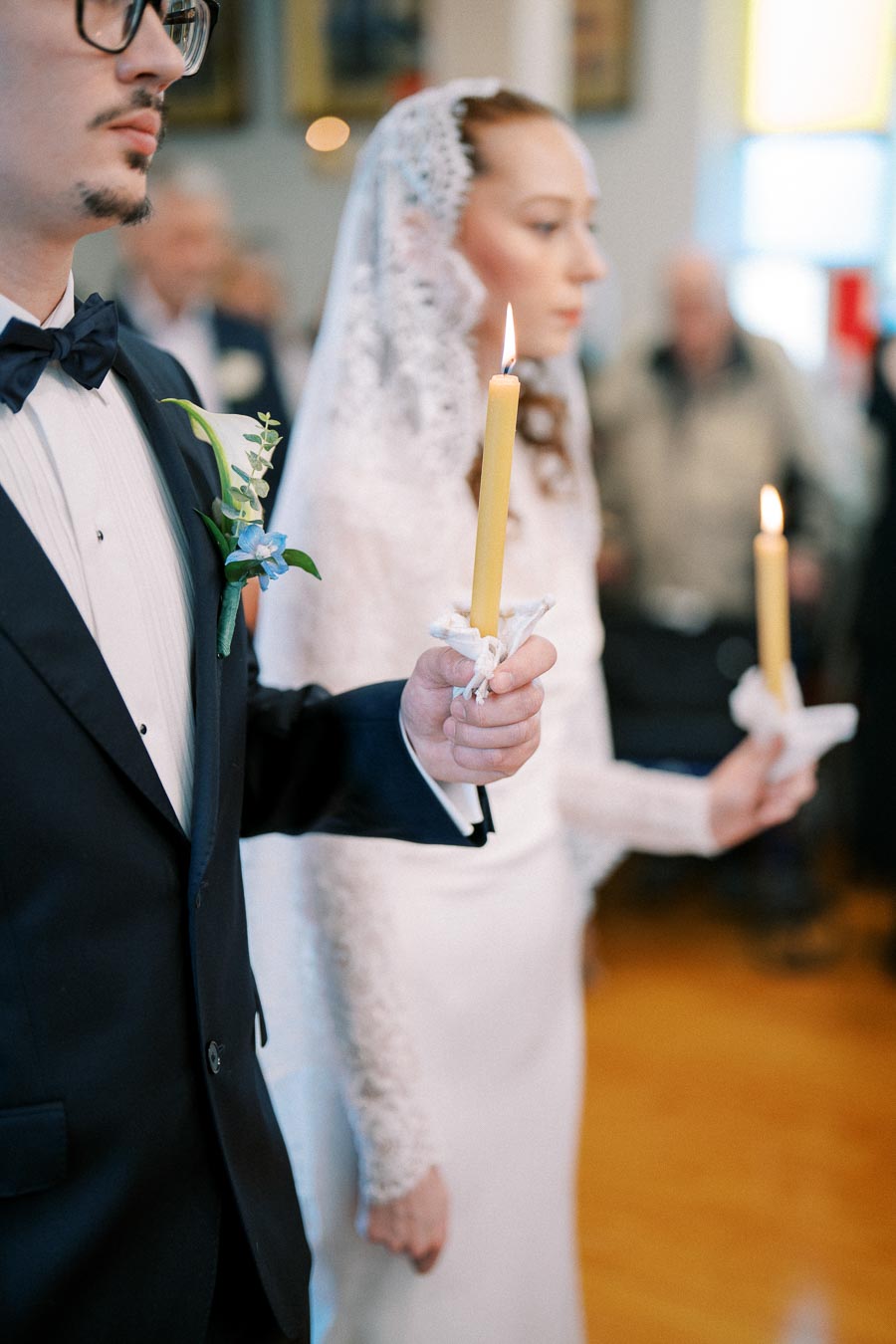 Bride and groom holding candles during an elegant wedding ceremony in a church, featuring a close-up of their hands and clothing details.
