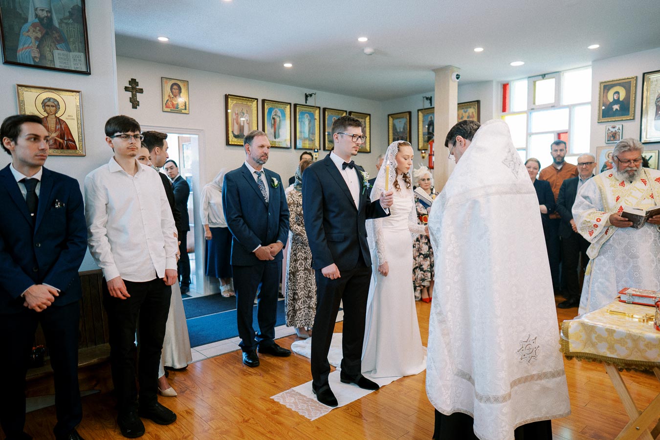 A traditional wedding ceremony takes place in an ornate church, featuring a couple in formal attire surrounded by guests and priests in religious garments, with religious icons displayed on the walls.