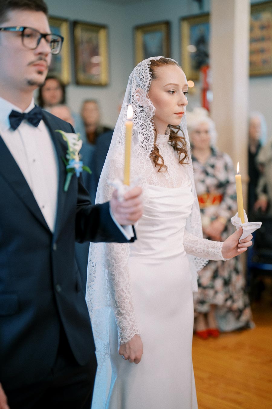 A bride and groom hold lit candles during a traditional wedding ceremony. The bride is wearing a lace gown with a veil, while the groom is dressed in a suit with a bow tie. They stand in a decorated venue with guests in the background.