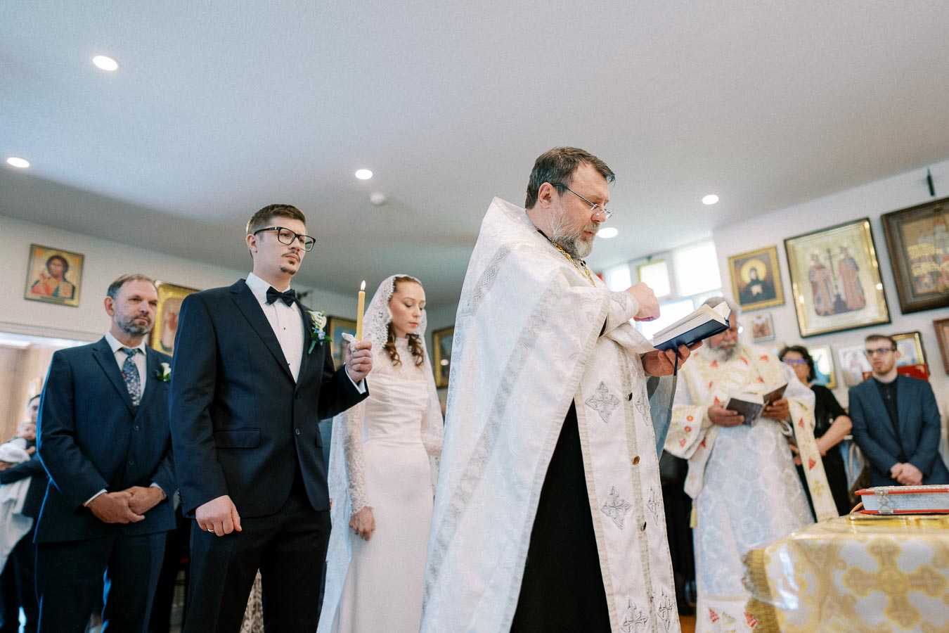 Orthodox wedding ceremony with bride and groom standing beside a priest holding a book, surrounded by religious icons on the walls.