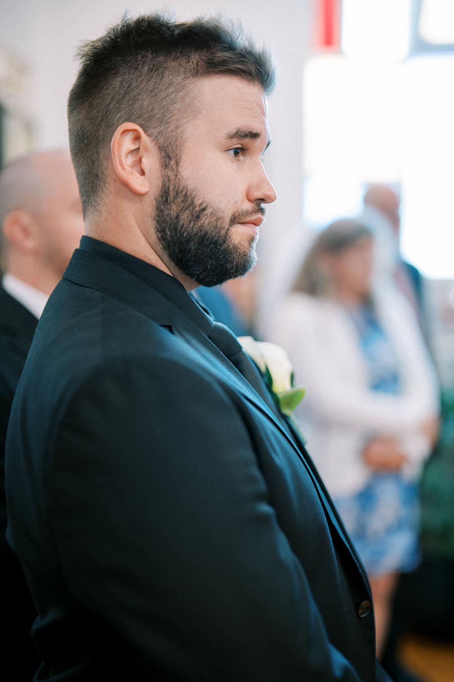 A man in a formal black suit with a white rose boutonniere stands in profile at a wedding ceremony, surrounded by softly blurred guests in the background.