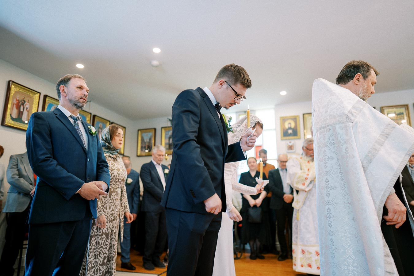 A couple participates in a traditional wedding ceremony, holding candles, surrounded by family and guests in formal attire. The room is adorned with religious icons on the walls.