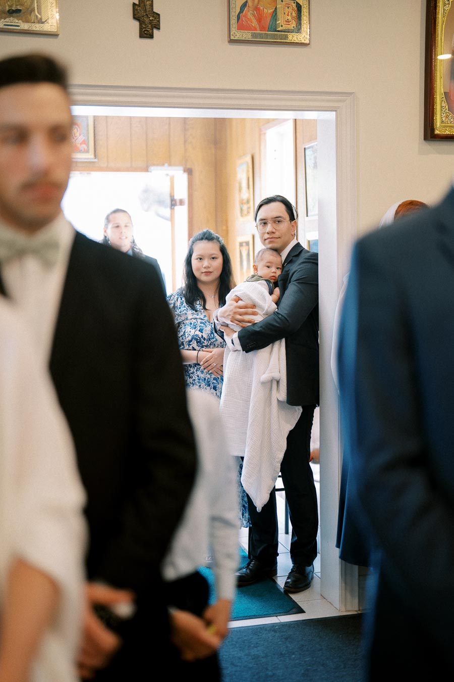 Family holding a baby in a church setting during a social gathering or event, with people in formal attire in the foreground.