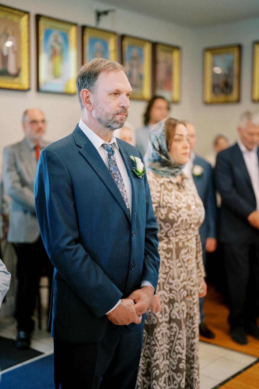 A man in a blue suit stands next to a woman in a patterned dress during a formal ceremony in a room adorned with framed art.