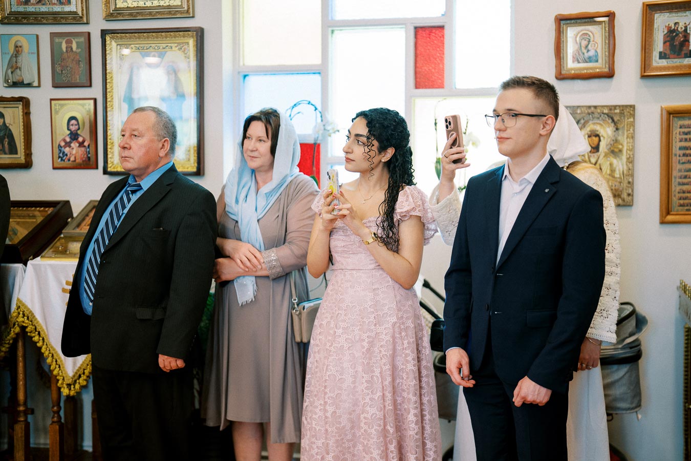 A group of people standing inside a room with religious icons on the walls, dressed in formal attire, some holding smartphones.