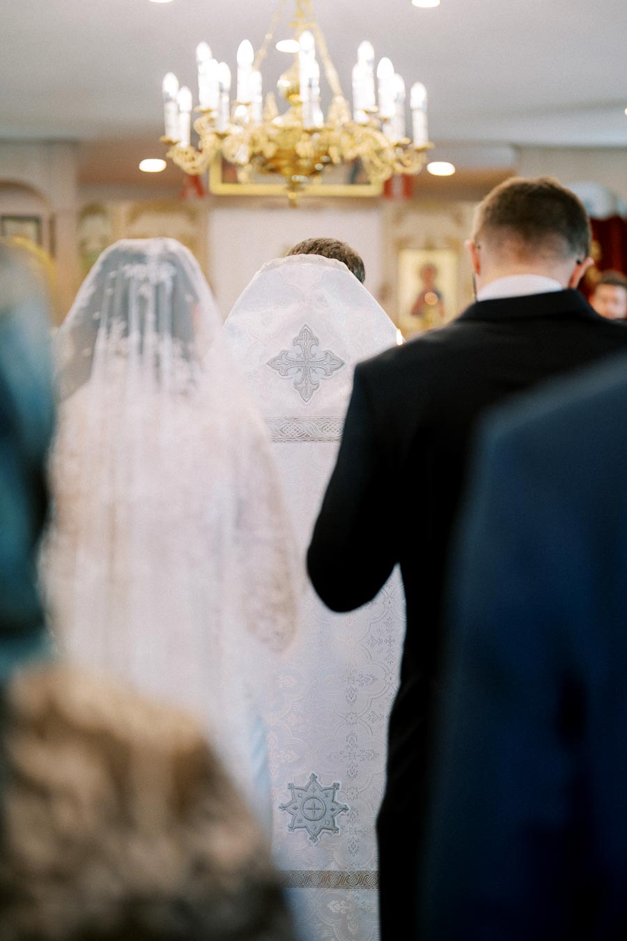 Traditional wedding ceremony with bride and groom in a church, featuring ornate details on wedding attire and chandelier in the background.