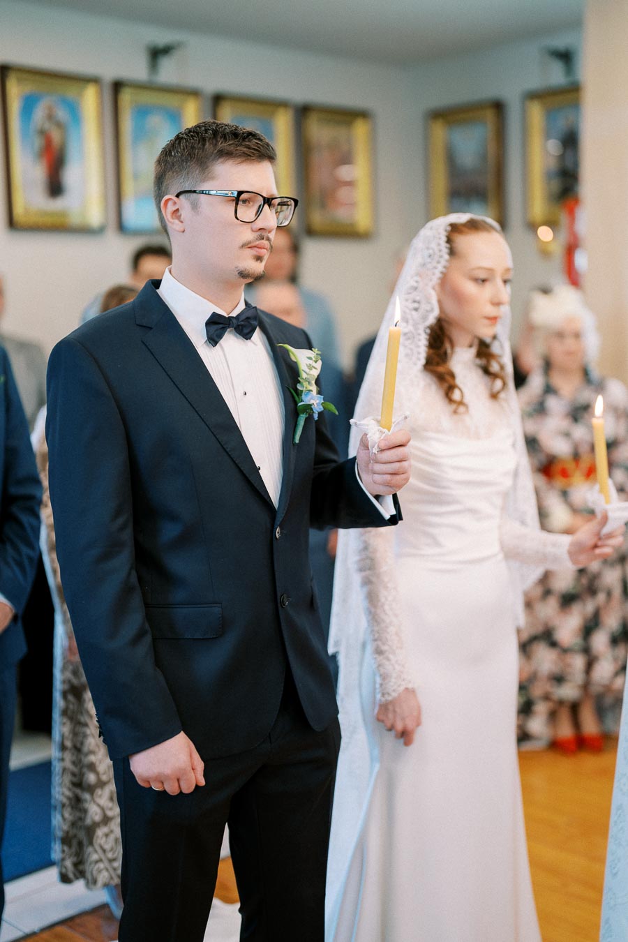 A bride and groom hold candles during a traditional wedding ceremony, wearing a black tuxedo and a white bridal gown with a lace veil.