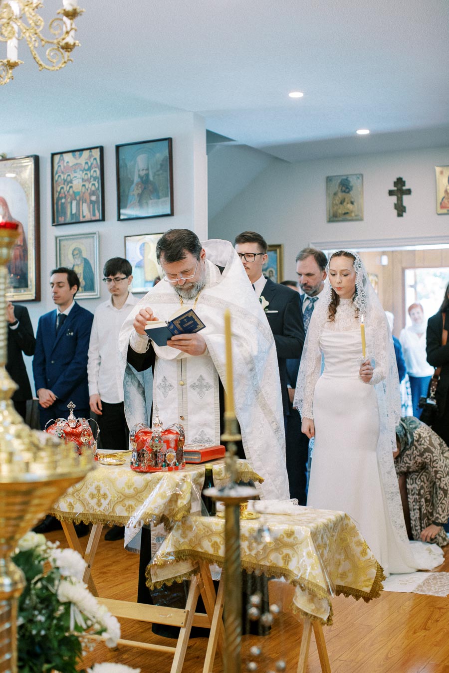 A priest conducting a traditional Orthodox wedding ceremony inside a decorated church, with a bride in a white dress holding a candle and guests observing.