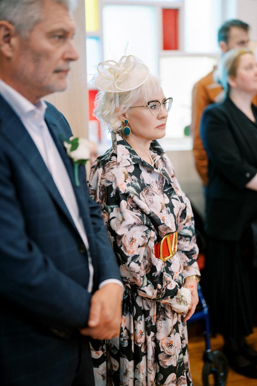 Elegant woman in floral dress and fascinator attending event, standing beside man in suit.