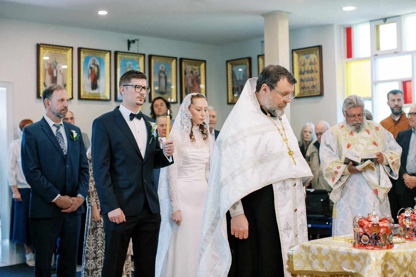 Orthodox wedding ceremony with bride and groom in traditional attire, standing beside a priest wearing a white robe, surrounded by guests in a decorated church setting.