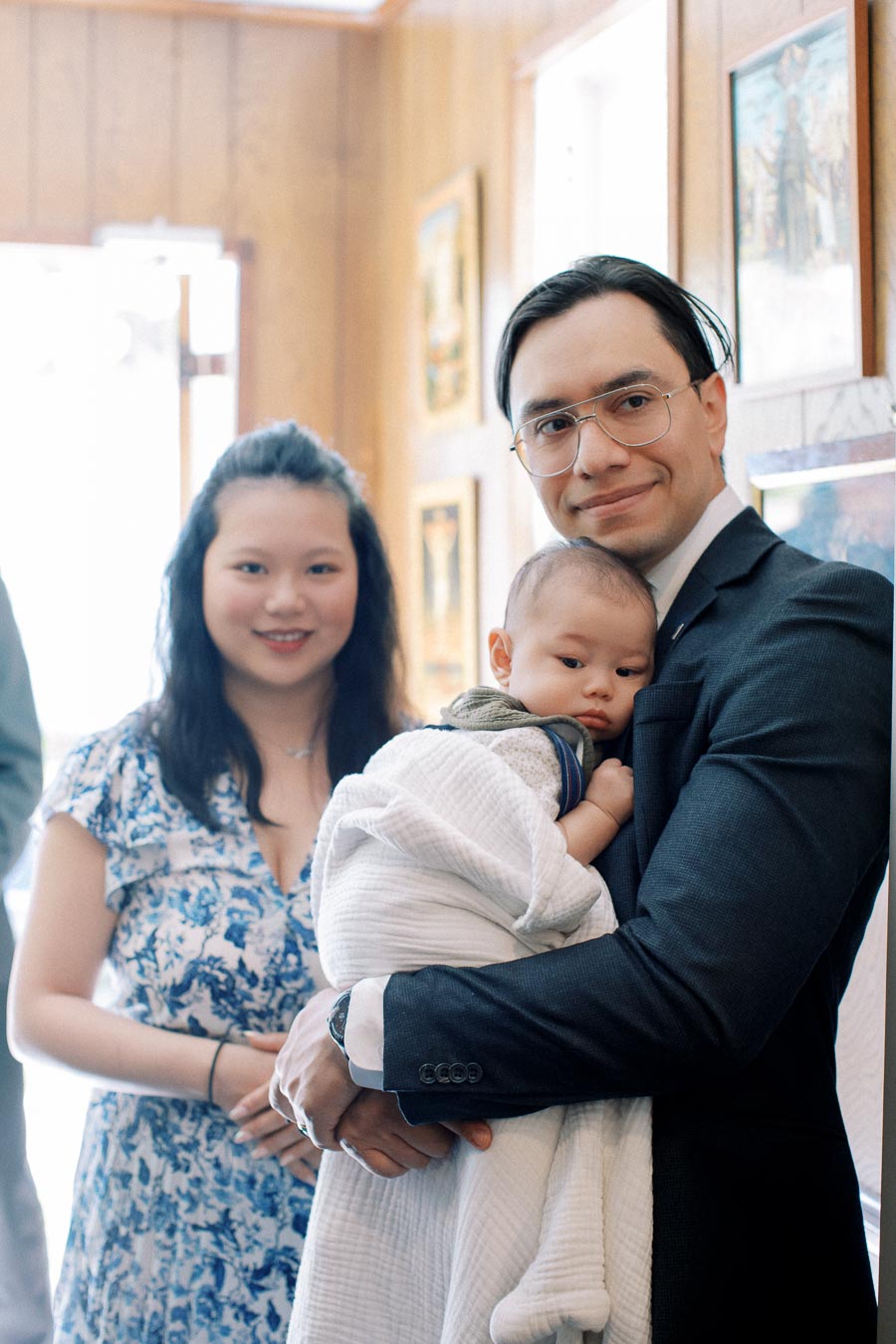 Smiling man in suit holding a baby wrapped in a white blanket, with a woman in a floral dress standing behind, inside a warmly decorated room with framed artwork on the walls.