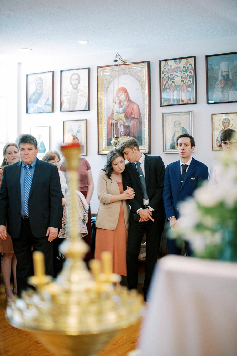 A group of people stands solemnly inside a church, surrounded by religious icons and wall decorations, creating a serene and contemplative atmosphere.