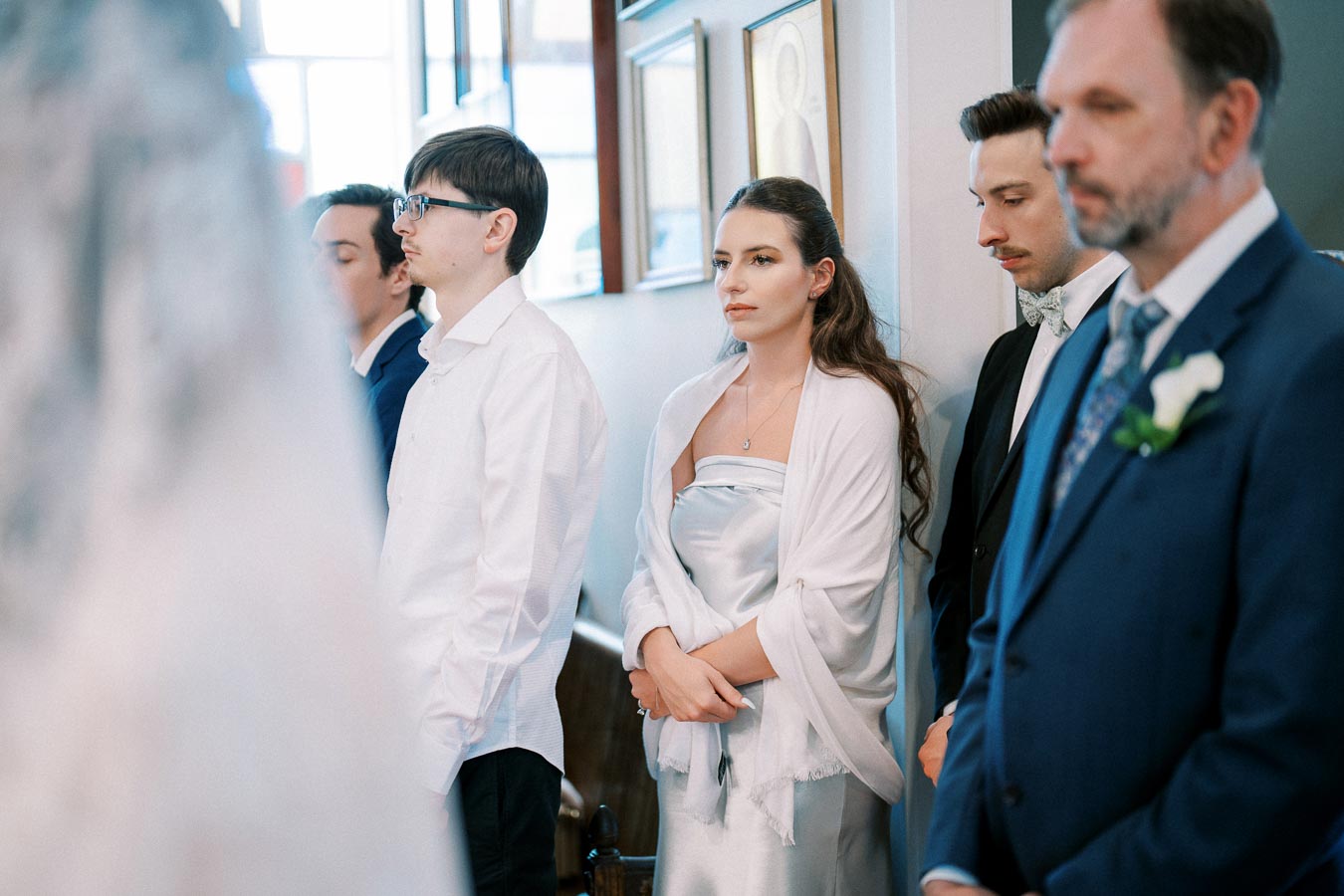 A group of elegantly dressed guests standing attentively at an indoor wedding ceremony, with a focus on a woman in a silver dress and white shawl.