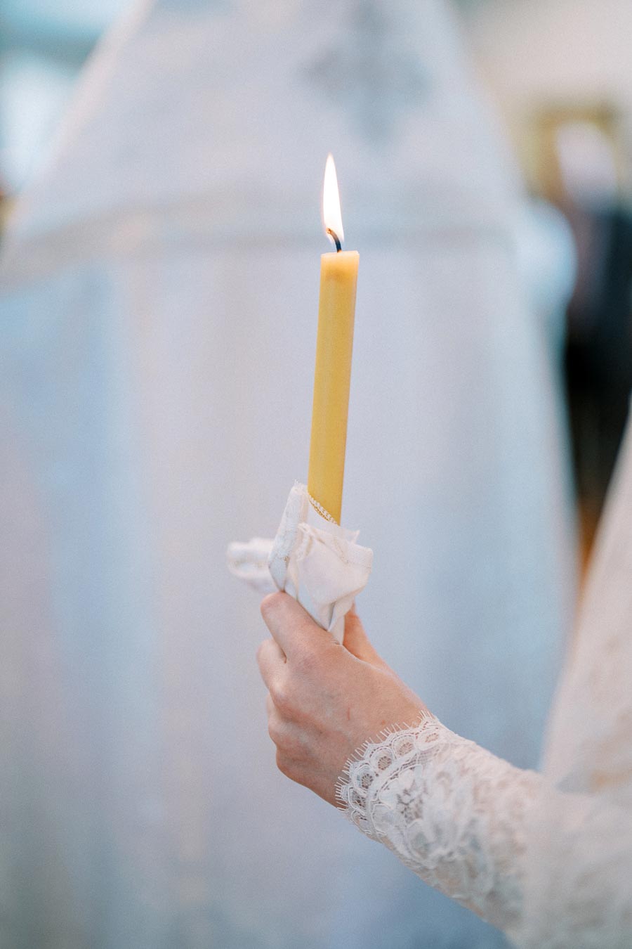 A person holding a lit candle wrapped in a white cloth, wearing a delicate lace sleeve, symbolizing purity and spirituality in a serene setting.