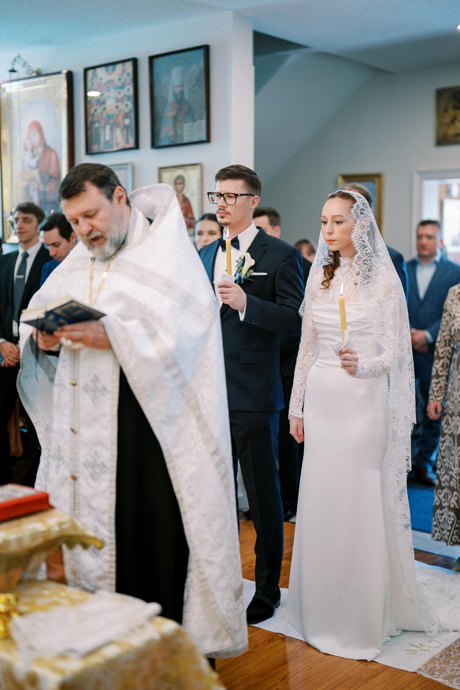 Ceremony in Orthodox church with priest leading a wedding procession. Groom and bride, in formal attire, hold candles while standing together.