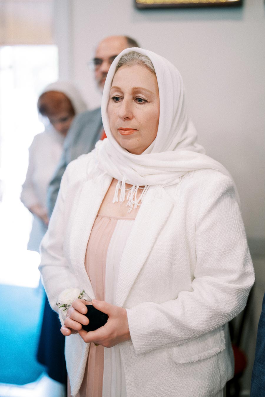 Woman in a white headscarf and jacket holds a small black object, standing in a softly lit room with blurred background.