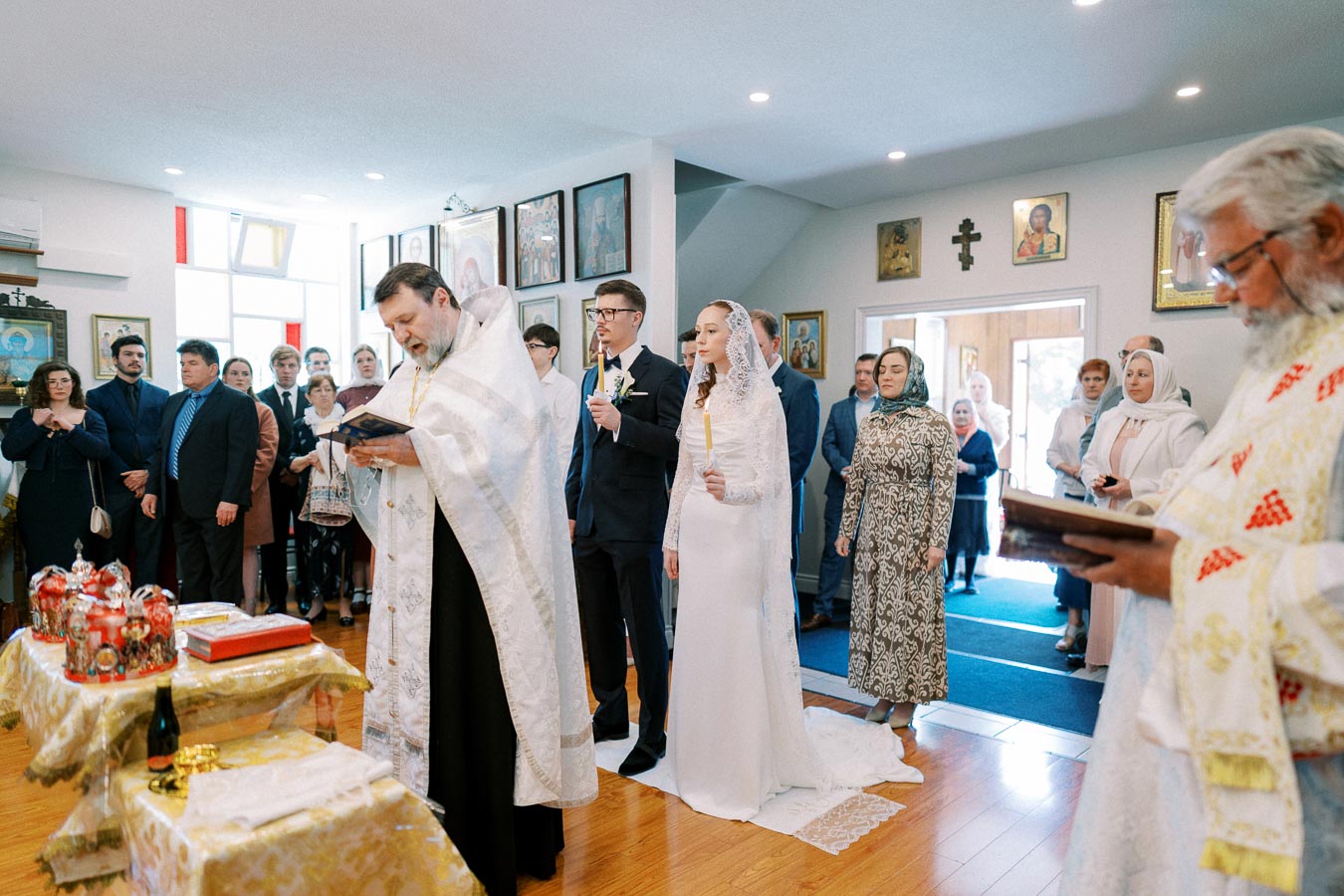 Orthodox wedding ceremony in a church with a priest leading the rituals, the bride in a white gown and veil, the groom holding a candle, and guests in formal attire observing the ceremony surrounded by religious icons.