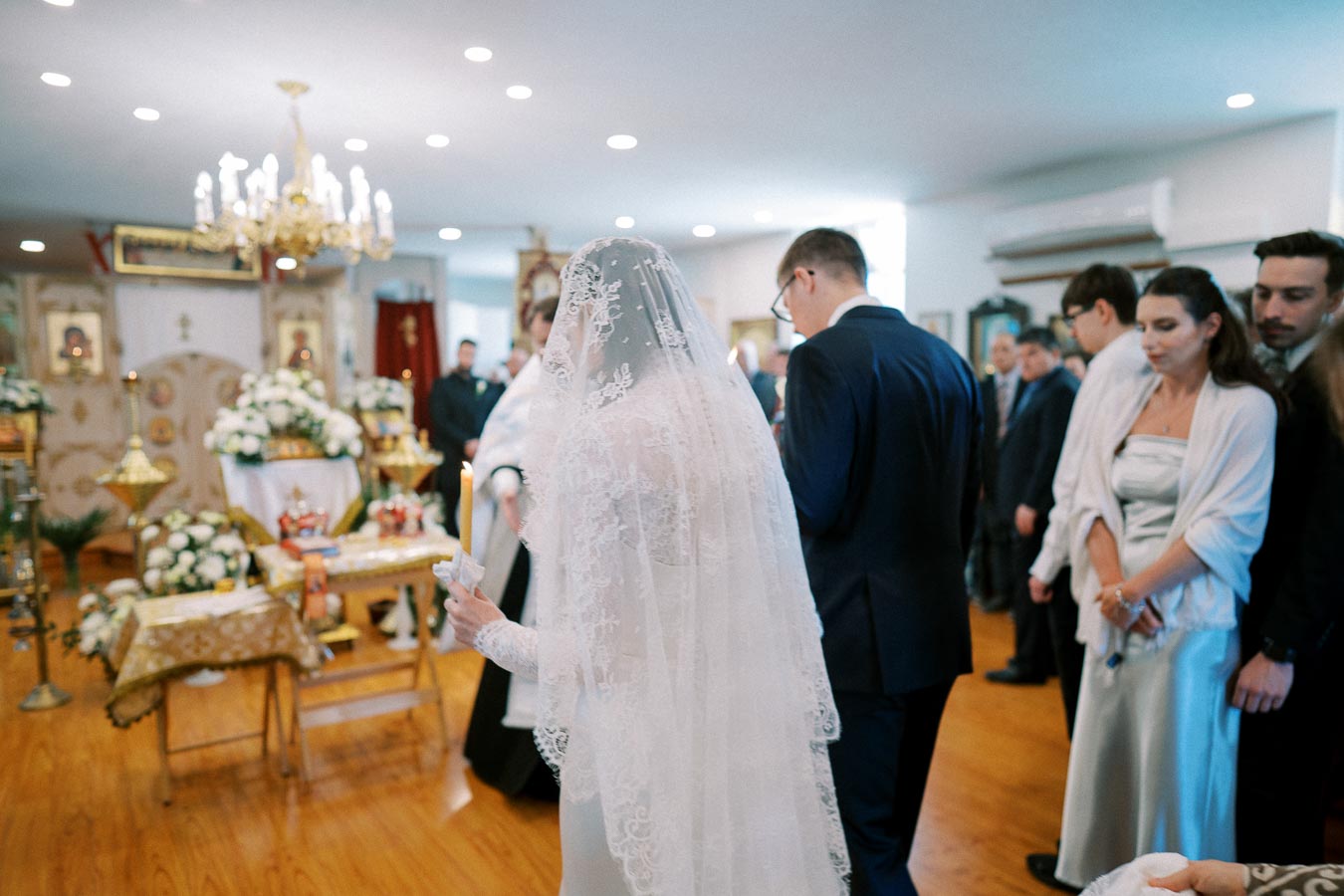 A bride in a lace veil holding a candle during a traditional religious wedding ceremony in a beautifully decorated church interior, surrounded by guests and golden chandeliers.