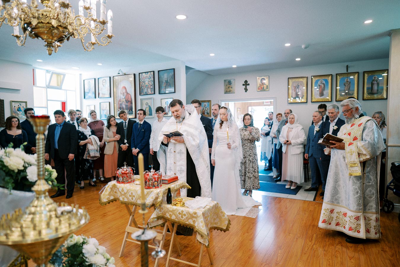 A traditional religious ceremony taking place in a decorated church interior. A priest in white robes reads from a book while standing beside a bride in a white gown holding a candle. Guests in formal attire observe, surrounded by religious icons and floral arrangements.