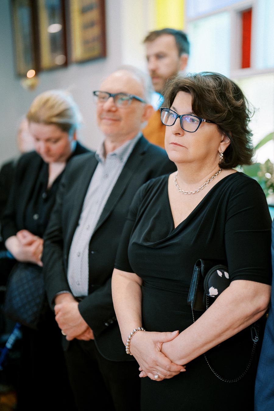 A group of people attentively listening during an indoor event, with a focus on a woman wearing a black dress and glasses, expressing deep concentration.