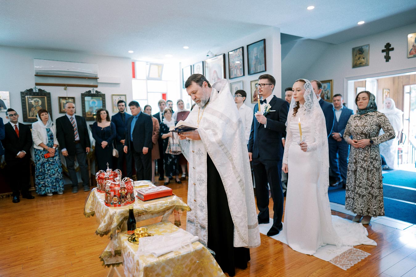 Orthodox wedding ceremony in a church, featuring a priest reading from a book, a bride and groom holding candles, and guests gathered around. The room is adorned with religious icons and traditional decor.