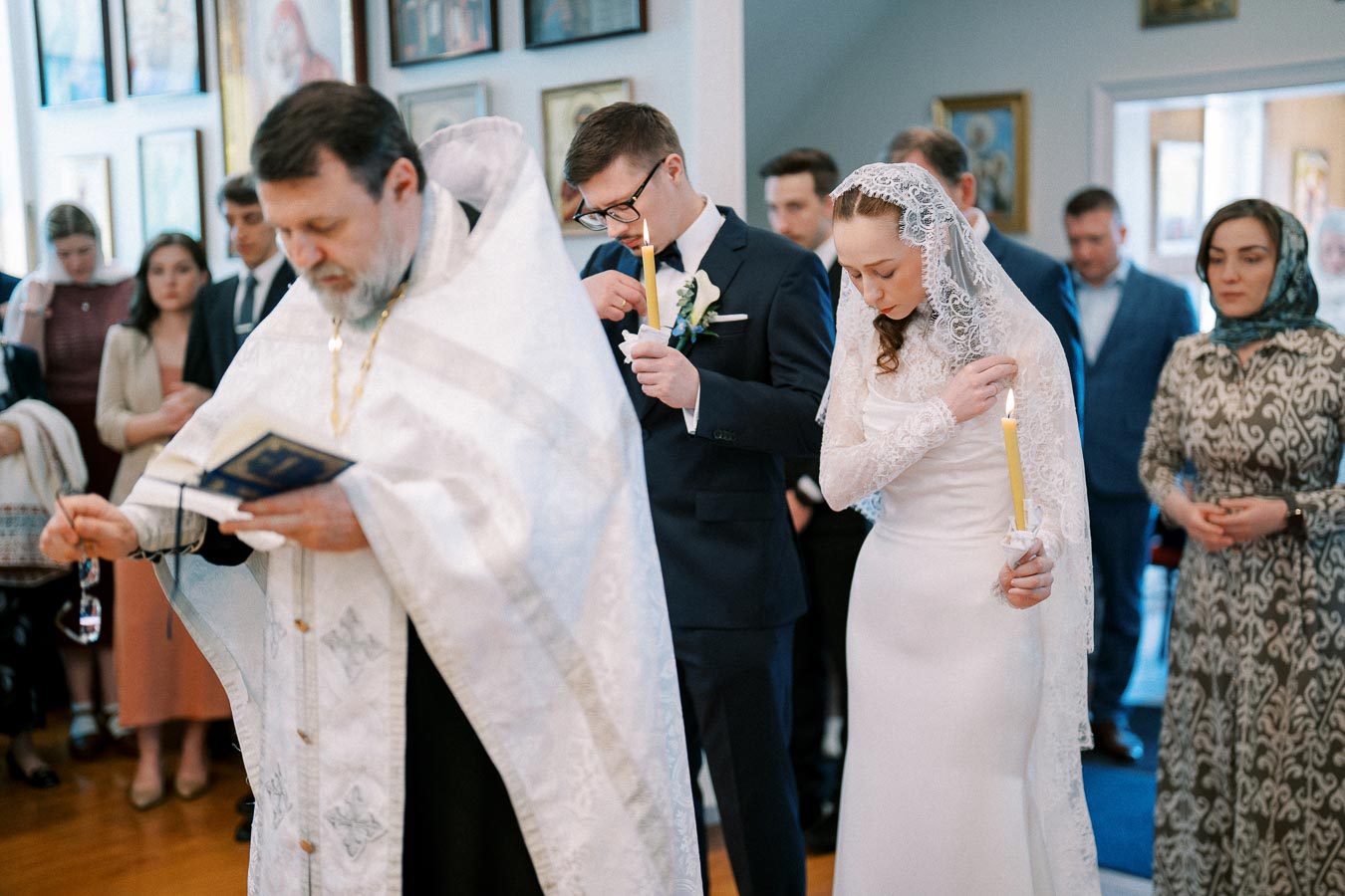 Orthodox wedding ceremony with a bride in a lace veil and groom holding candles, led by a priest in traditional attire, surrounded by guests inside a church with religious icons on the walls.