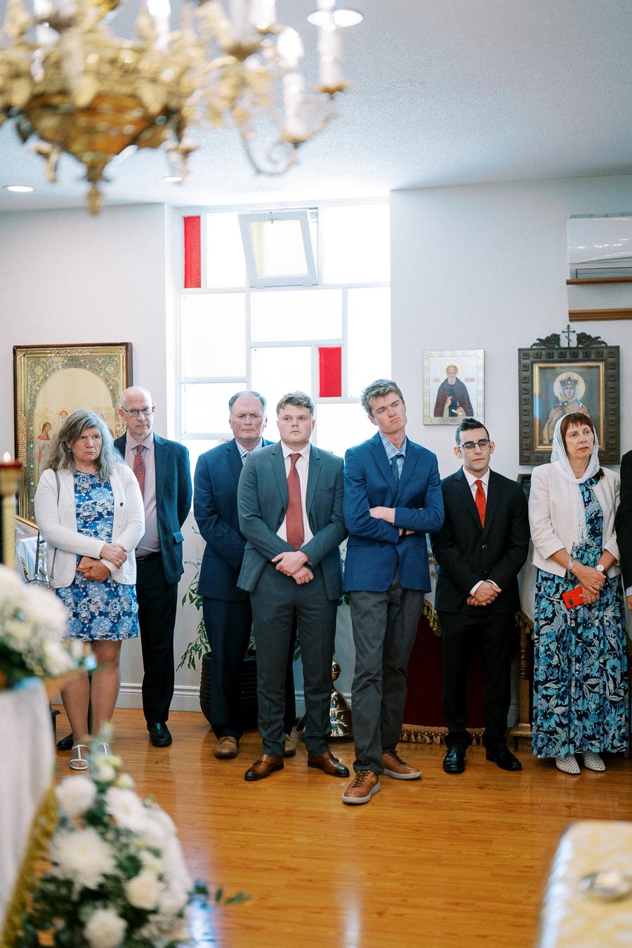 A group of people standing attentively in a decorated room, surrounded by religious icons and floral arrangements. They are dressed in formal attire, indicating a ceremonial or significant event.