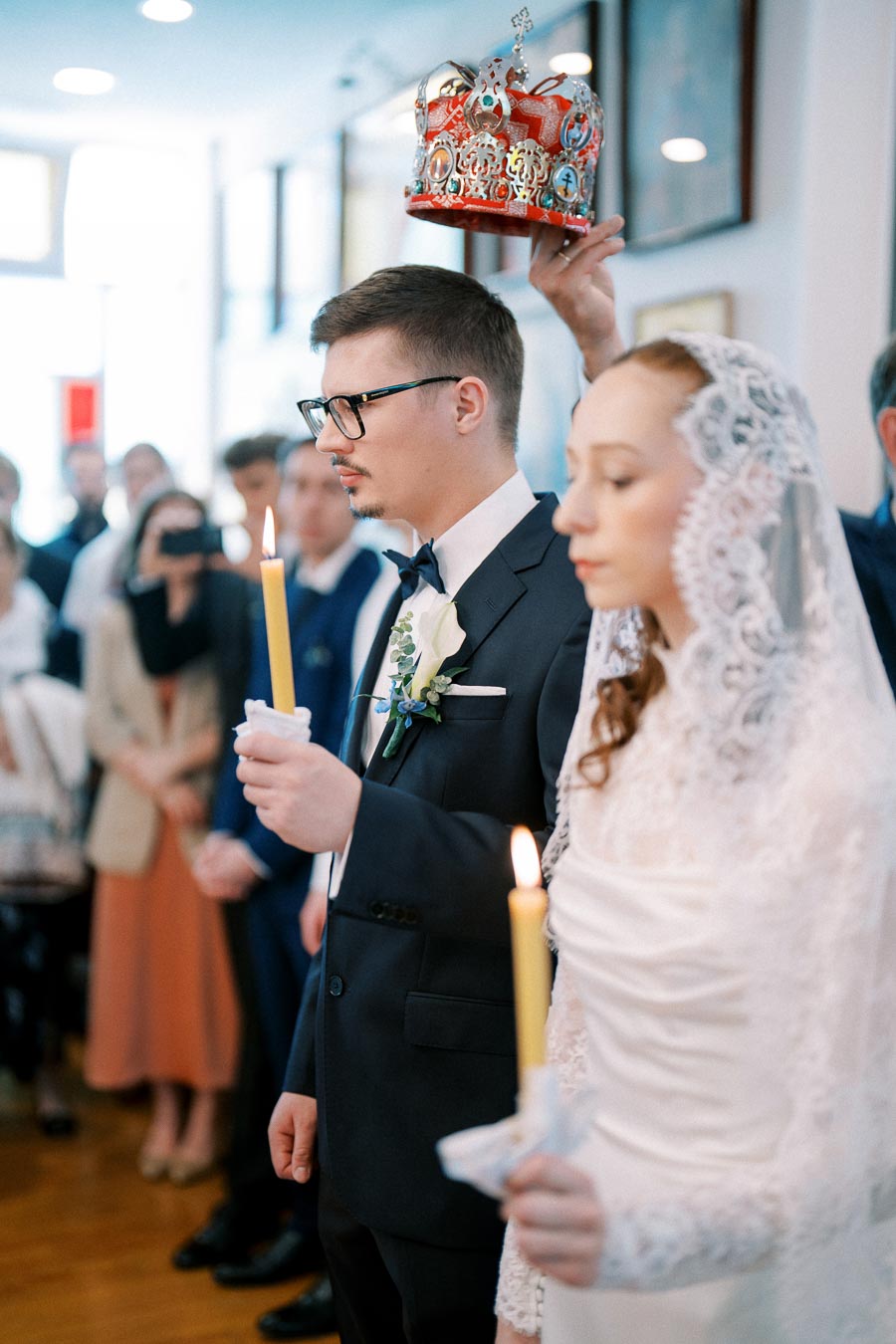 A bride and groom during a traditional wedding ceremony, each holding lit candles, with the groom wearing a suit and a floral boutonniere, and the bride in a lace-covered dress, while a crown is held above their heads indoors.