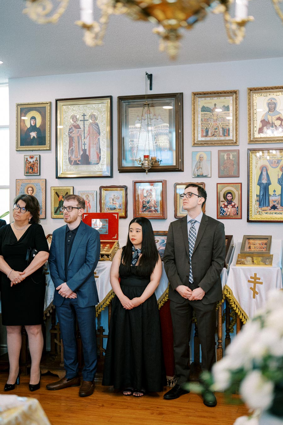 A group of people standing in an Orthodox Christian church, surrounded by religious icons and framed artwork, during a ceremonial event or service.