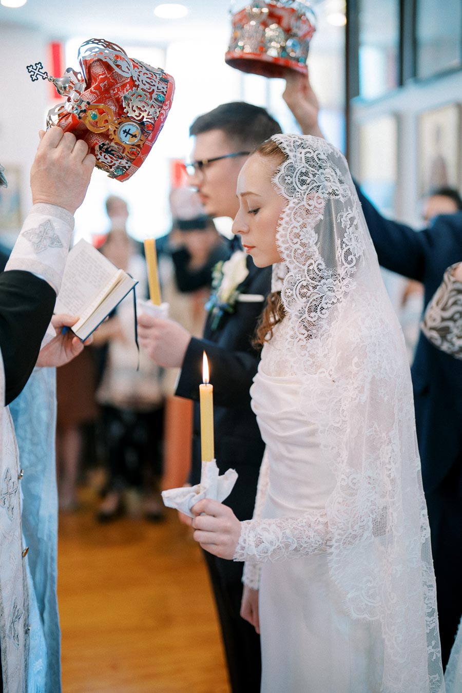 Bride and groom participate in a traditional wedding ceremony, adorned in intricate attire, holding lit candles as crowns are placed above their heads by an officiant.