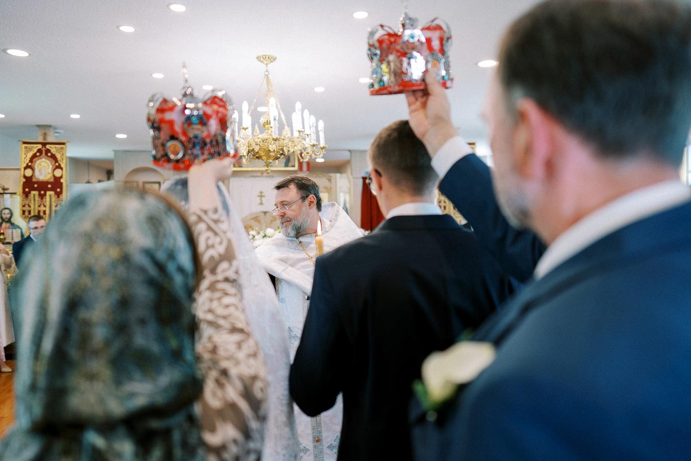Orthodox wedding ceremony with priest leading rituals as men hold ornate crowns, illuminated by a golden chandelier in a church setting.