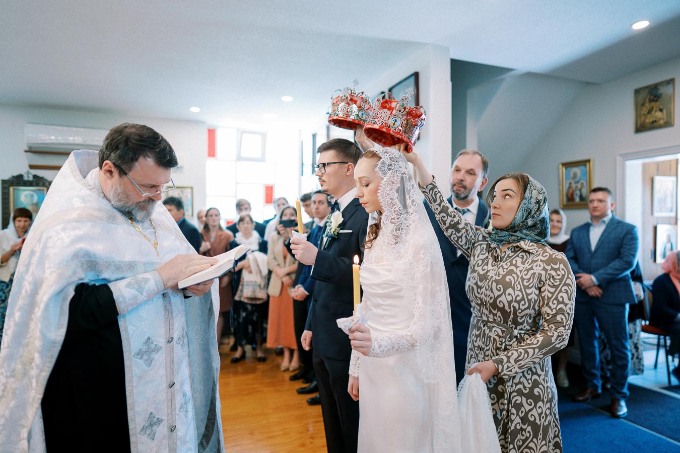Orthodox wedding ceremony inside a church, featuring a bride and groom holding candles, surrounded by a priest and guests; crowns being placed on their heads as part of the traditional ritual.