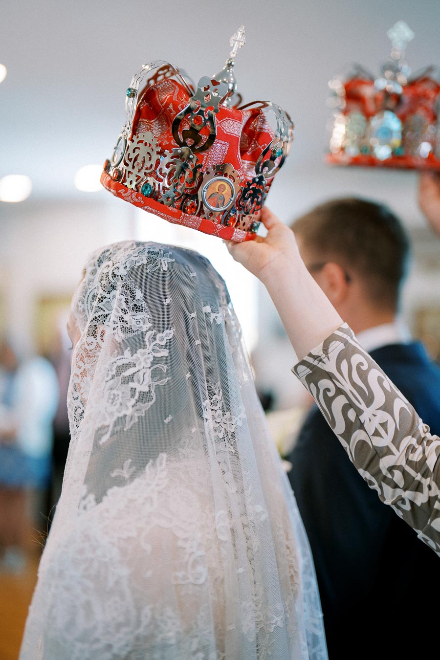 A person places an ornate red and silver crown over a bride wearing a lace veil at a traditional wedding ceremony.