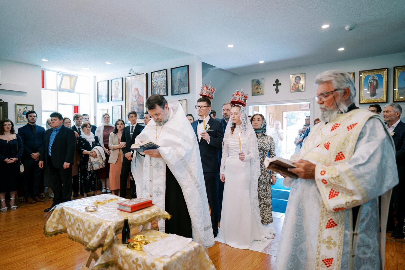 Orthodox wedding ceremony depicting a bride and groom wearing crowns, accompanied by priests in traditional vestments reading religious texts, surrounded by guests in a decorated church interior.