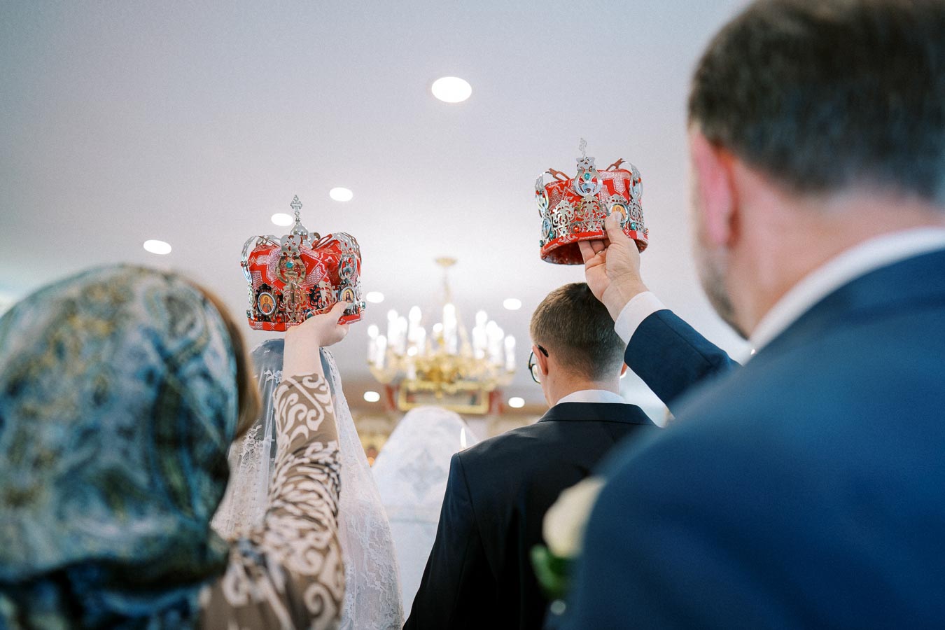 Ceremony participants holding decorative crowns above a bride and groom during an ornate wedding ritual inside a church, with a chandelier in the background.