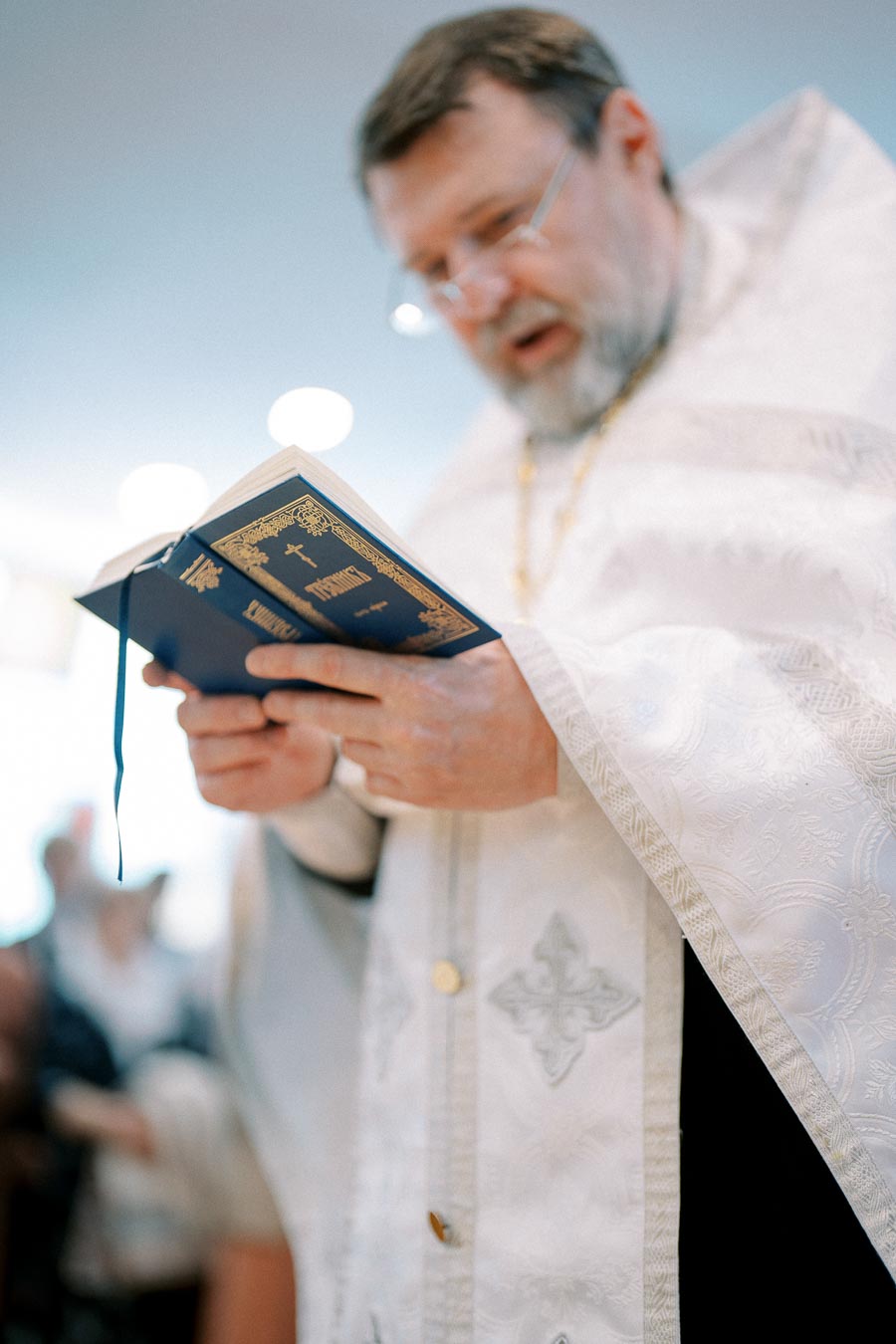 Priest reading from a religious book in a church ceremony, wearing ornate white vestments.
