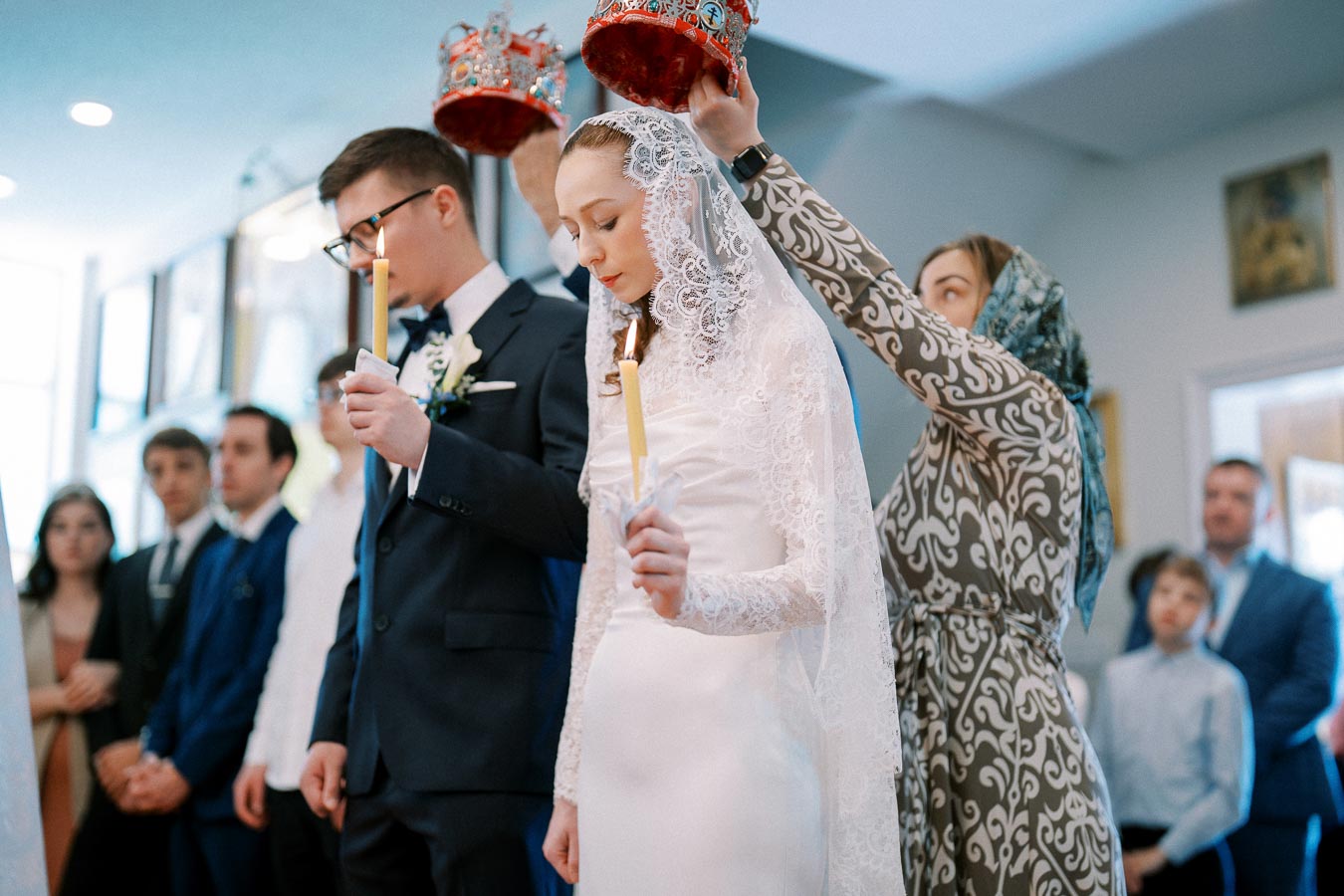 A couple standing in a traditional wedding ceremony, holding lit candles, with decorative crowns being placed on their heads by attendants. The bride is wearing a lace veil and the groom is in a suit with a white flower boutonniere. Celebratory scene in a church setting.