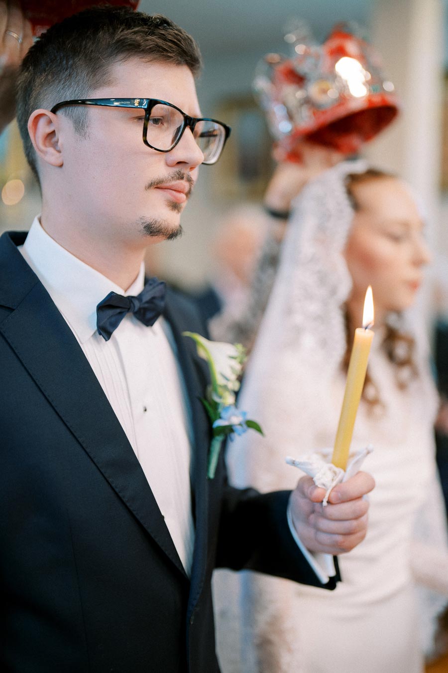 Man in formal suit holding a candle during a wedding ceremony, with crown being placed on his head. Bride in background, wearing a veil, also holds a candle.