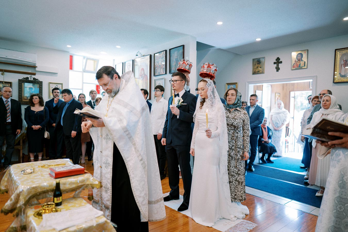 Orthodox wedding ceremony with priest blessing couple, bride holding candle, and guests observing in decorated church.
