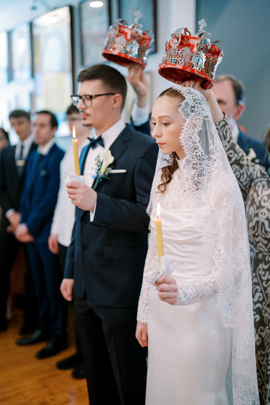 A bride and groom standing during an Orthodox wedding ceremony, holding candles with crowns being placed on their heads, surrounded by wedding guests.