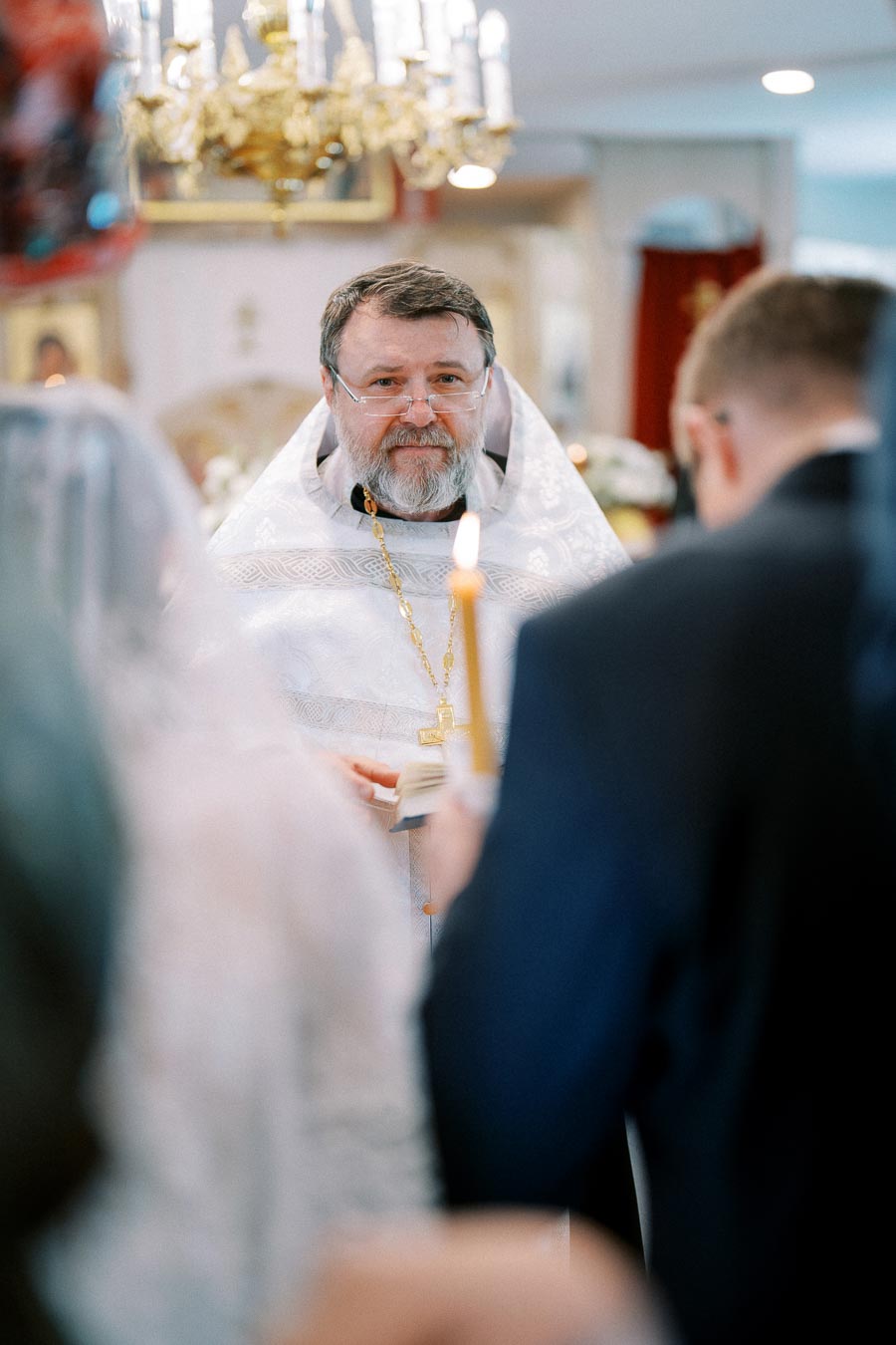 A priest in white liturgical attire stands in a church, holding a religious book during a ceremony. Attendees in formal clothing are visible, and the warm light of a candle enhances the serene atmosphere.