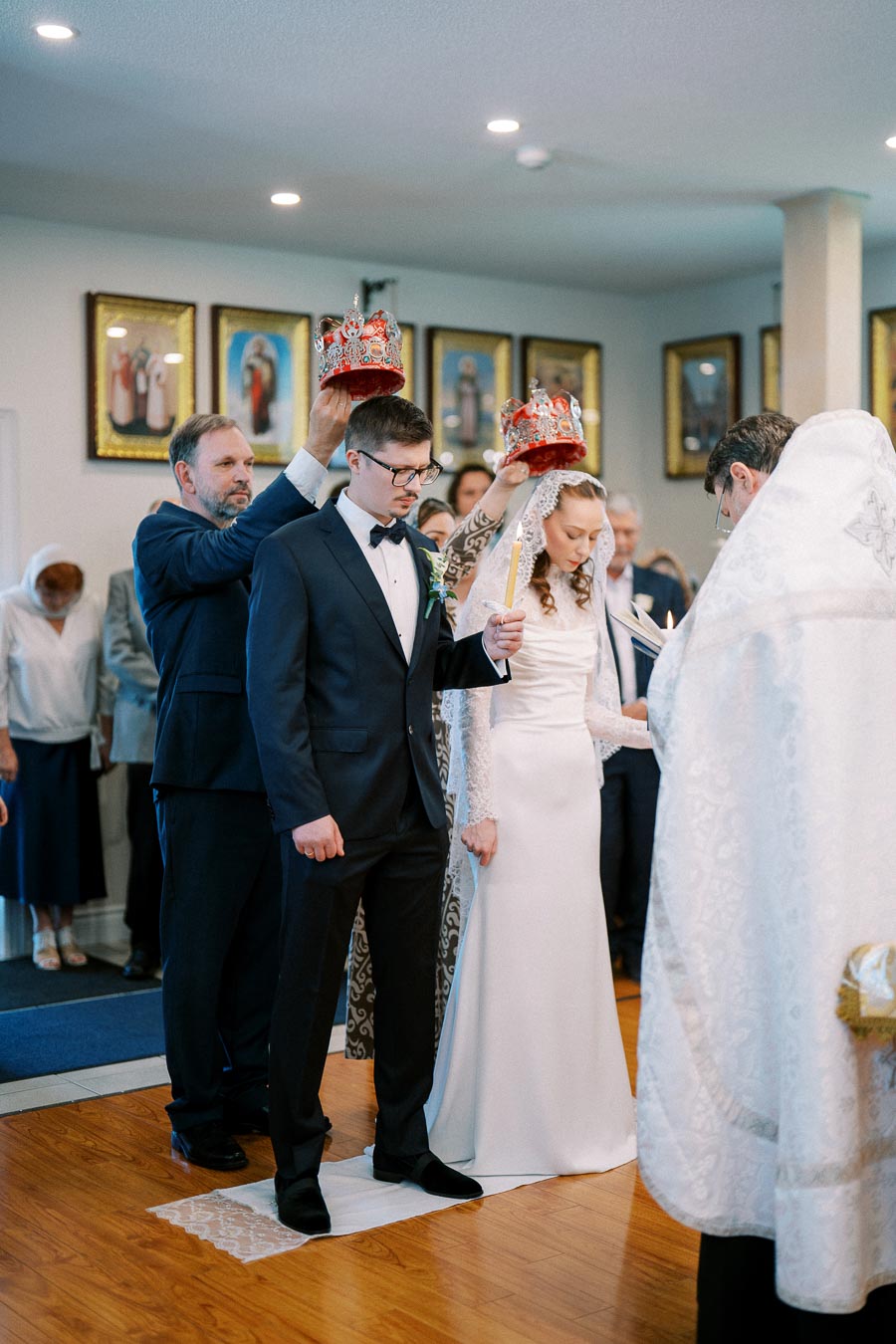 Orthodox wedding ceremony with a bride in a white lace dress and groom in a suit, each holding a candle. Crowns are being placed on their heads by attendants. The ceremony is taking place in a church with religious icons on the walls and guests in the background.