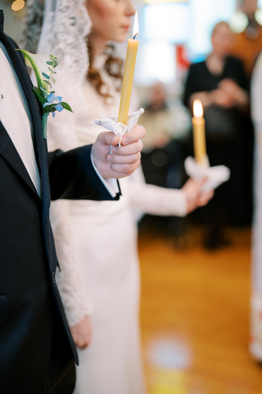 A couple holding lit candles during a traditional wedding ceremony, with the bride wearing a lace gown and veil, and the groom in a formal suit decorated with a blue flower boutonniere.