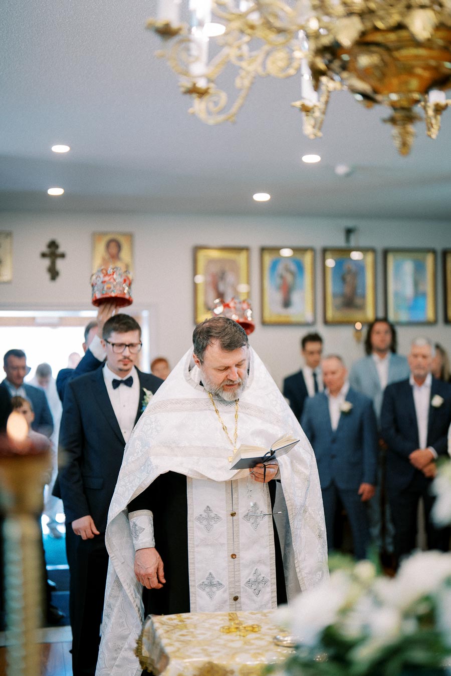 Orthodox wedding ceremony in church with priest reading from a book, groom in a navy suit, ornate crowns held over heads, framed religious icons on the wall, floral decorations, and golden chandelier in foreground.