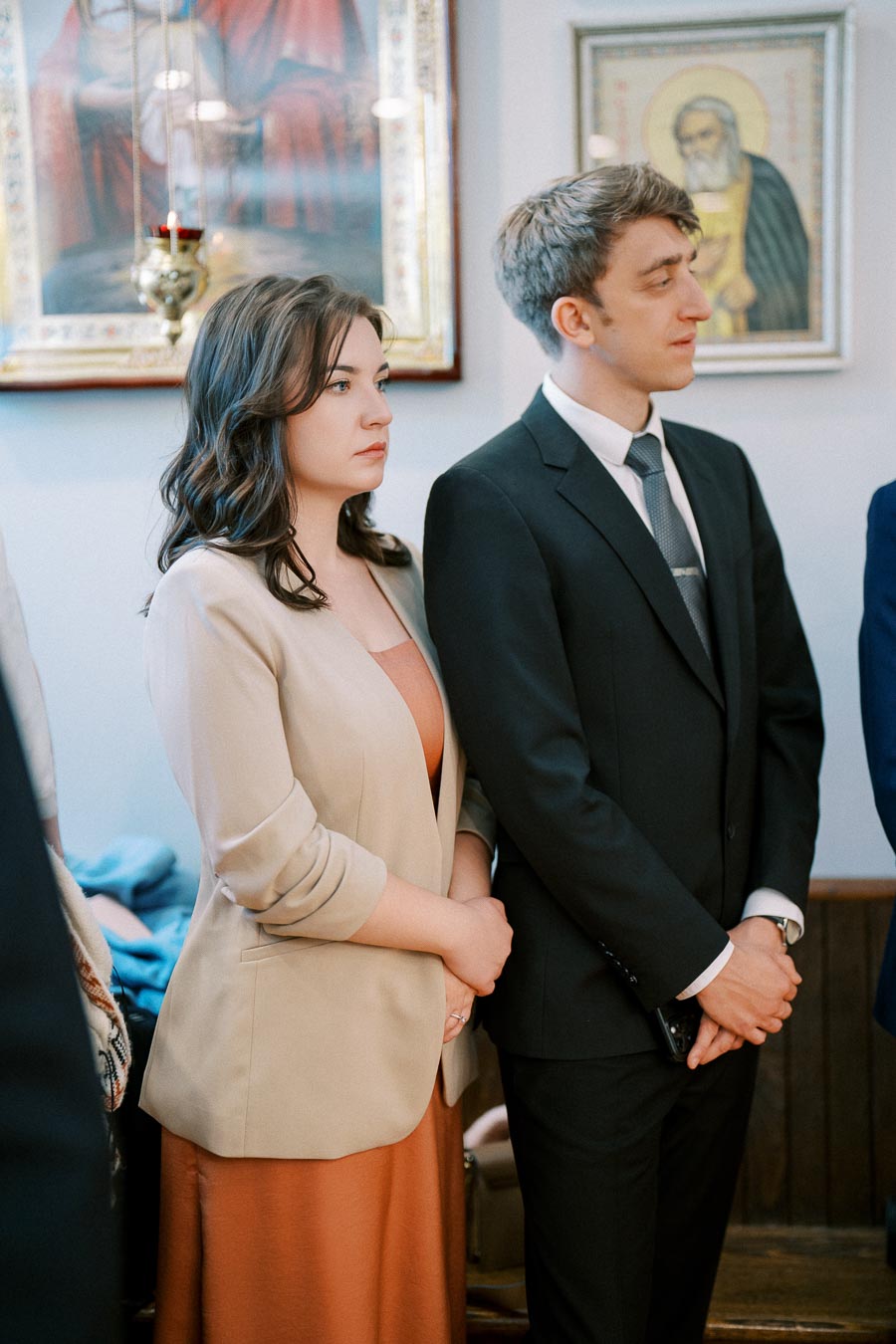 A man and woman in formal attire standing side by side in an art gallery, with framed religious artwork in the background.