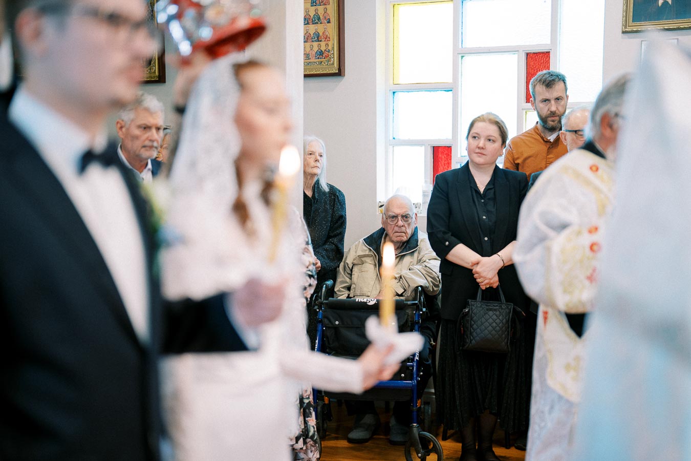 A wedding ceremony inside a church, featuring a bride holding a candle in focus, with blurred guests and a priest in the background.