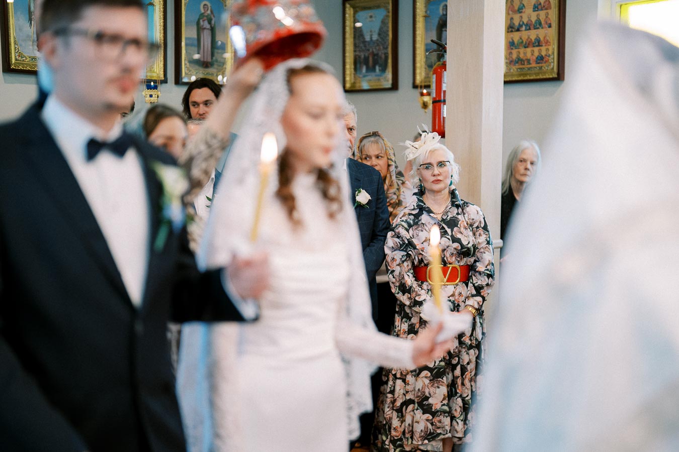 A wedding ceremony in a church, with a bride and groom holding candles in the foreground. Guests are watching attentively, with ornate religious icons visible on the walls behind them.